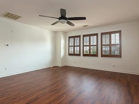 A wide, empty living room with light grey walls and light wood flooring. A large window is visible on the far wall, letting in natural light.